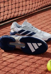 Chaussures de tennis blanches avec une semelle texturée à base bleue, trois bandes bleu marine et des accents gris clair. Placées près d'une balle de tennis sur terre battue.