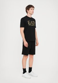Black cotton t-shirt with gold "EA7 Emporio Armani" logo, paired with black shorts. Model wears white sneakers and black crew socks.