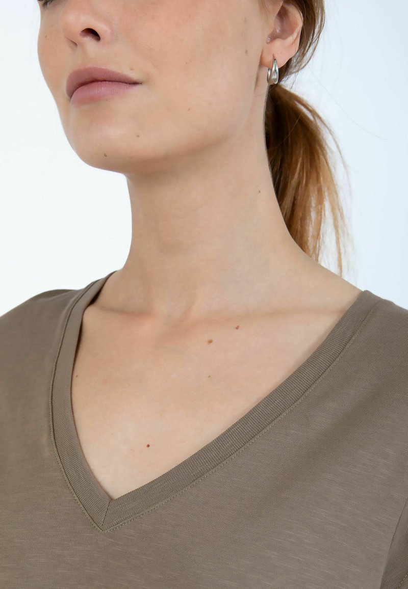 Olivengrøn V-hals skjorte med en glat tekstur. Har en løs pasform og synlige syninger omkring halsudskæringen. Enkel design, uden mønstre.