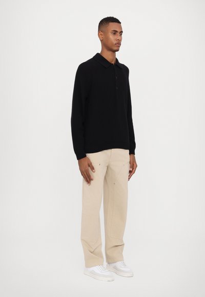 Polo en tricot noir avec un col, quatre boutons et des manches longues, associé à un pantalon beige clair et des baskets blanches, se tenant sur un fond uni.