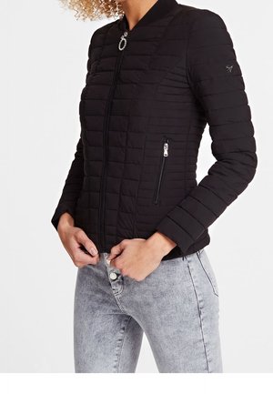 Vrouw draagt een aansluitend zwart doorgestikt jack met ritszakken en lichtgrijze spijkerbroek, handen nabij de rits van het jack.