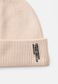 Gorro de punto acanalado beige con puño doblado, que presenta una etiqueta de logo negra en el costado. Textura suave y diseño ajustado.