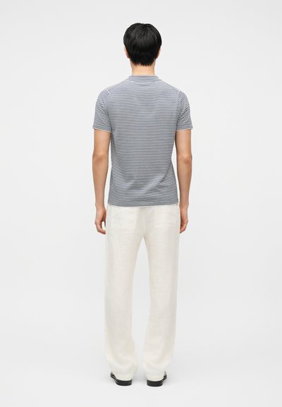 Homme debout, de dos, portant un T-shirt à manches courtes rayé noir et blanc, un pantalon blanc et des chaussures noires sur un fond blanc.