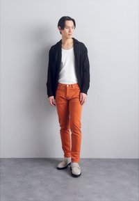 Svart stickad cardigan över en vit t-shirt, orange byxor och beige skor med svarta sulor. Enkla texturer och figurnära designelement.