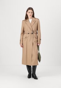 Bézs gyapjúpulóver anyagú trench coat, övvel a deréknál, vállvédőkkel és két elülső zsebbel. Fekete térdig érő csizmákkal és zöld táskával viselve.