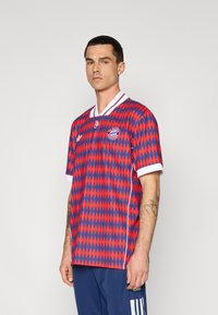 Czerwono-niebieski, wzorzysty dres piłkarski z diamentowymi kształtami, biały kołnierzyk i logo na piersi. Krótkie rękawy i lekki materiał.