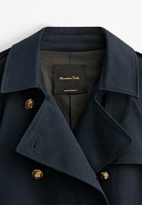 Trench-coat bleu marine avec larges revers, boutons décoratifs dorés et une étiquette noire indiquant « Massimo Dutti Made in Portugal » à l'intérieur du col.