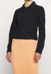 Svart denimjacka med knäppning, två bröstfickor och en kortad design. Tyget har en slät struktur som kompletterar en orange ribbad kjol.