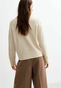 Beżowy sweter z dzianiny o luźnym kroju, z ribbowanym wykończeniem i długimi rękawami, połączony z brązowymi spodniami typu wide-leg. Miękka tekstura, minimalistyczny design.