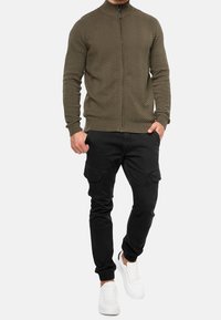 Olive green knitted zip-up jacket with a herringbone pattern, paired with black cargo pants and white sneakers.