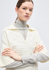 Jeune femme aux cheveux tirés en arrière, portant un col roulé gris sous un haut en maille texturée crème, les bras croisés, expression neutre, fond clair.