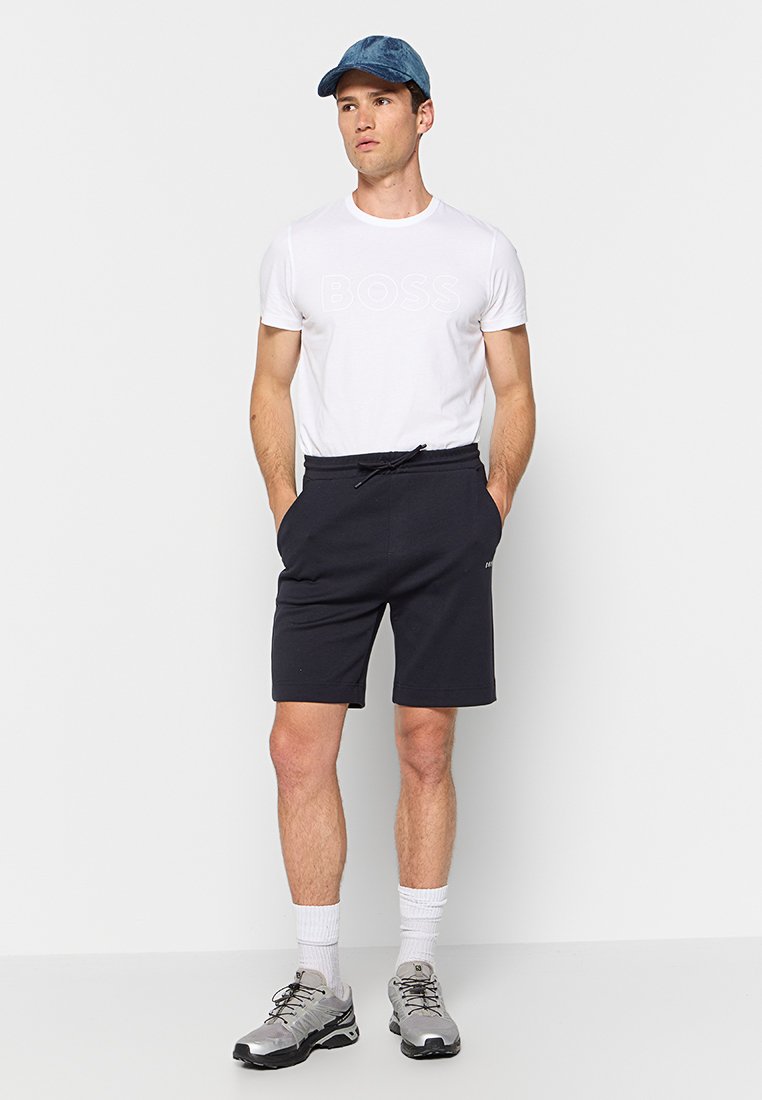 T-shirt blanche avec logo "BOSS" en relief, shorts noirs avec taille élastique, casquette bleue et chaussures de sport grises avec chaussettes blanches.