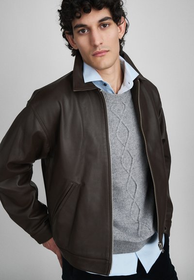 Braune Lederjacke mit Reißverschluss vorne, Seitentaschen, über einem grauen Strickpullover und einem hellblauen Hemd. Glatte Textur, modernes Design.