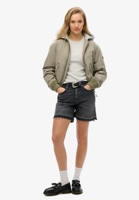 Olivengrøn bomberjakke med en grå hættetrøje, parret med højtaljede sorte denim-shorts. Sorte sko og hvide sokker fuldender outfittet.