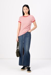 Gestreiftes rot-weißes T-Shirt mit kurzen Ärmeln, kombiniert mit einem langen blauen Jeansrock mit einem Schlitz vorne. Schwarze Sneaker und eine grüne Tasche.