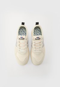 Baskets beiges avec un dessus en daim et textile, présentant une semelle intérieure à motif bleu, des lacets blancs et des accents en boucle noirs au niveau du talon.