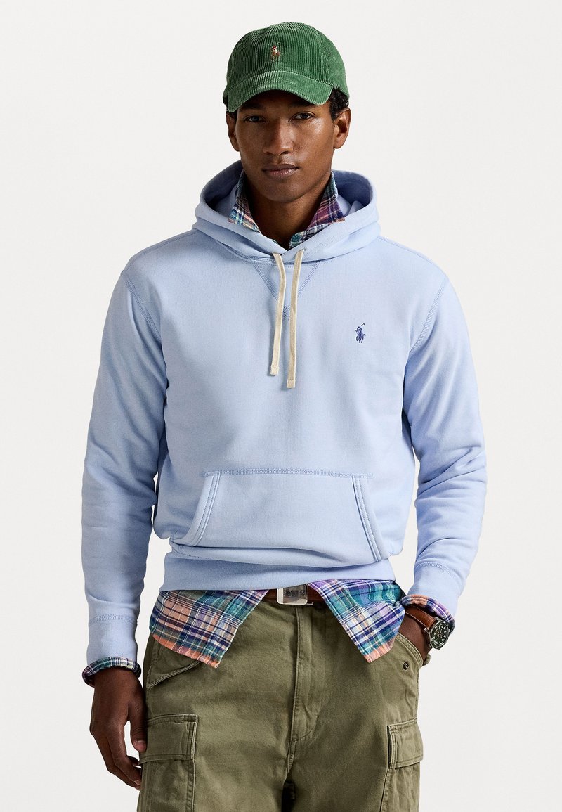Helle blaue Kapuzenjacke mit einer Fronttasche, cremefarbenen Kordeln und gesticktem Logo. getragen über einem mehrfarbigen karierten Hemd und olivgrünen Cargo-Hosen.