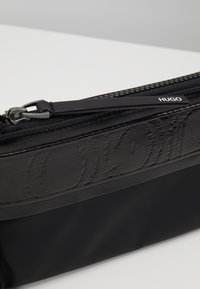 Pochette nera con zip realizzata in tessuto liscio con dettagli in rilievo del logo. Presenta una zip robusta e un design elegante e minimale.