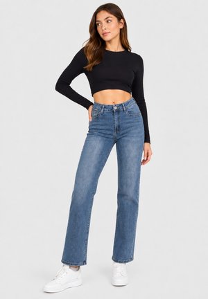 Vrouw draagt een zwarte crop top met lange mouwen, hooggetailleerde blauwe spijkerbroek en witte sneakers, staand met één hand op de heup.