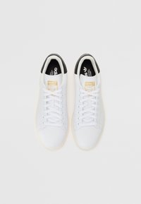 Sapatilhas brancas em couro com um acabamento liso, bico arredondado, atacadores planos e um detalhe em preto no calcanhar. Apresentam a marcação em dourado "Stan Smith".