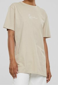 Camiseta de algodón beige con mangas cortas, cuello redondo y un logotipo bordado blanco en el pecho. Corte relajado y textura suave.