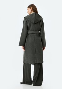 Lange grijze wollen jas met een capuchon, riem en een losse fit. Bevat een gestructureerde stof en wijde mouwen, gestyled met bijpassende grijze broek.