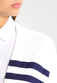Bílá bavlněná košile s límečkem, se námořnickomodra pruhovanou akcentací na rukávu. Hladká textura, detailní záběr, neutrální pozadí.