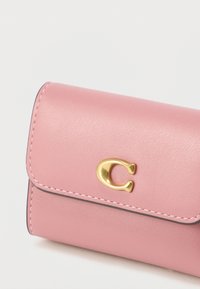 Ροζ δερμάτινο πορτοφόλι με καμπυλωτό καπάκι, χρυσή διακοσμητική λεπτομέρεια λογότυπου και αντίθετη ραφή κατά μήκος των άκρων. Συμπαγής ορθογώνιο σχήμα.