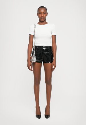 Femme portant un haut blanc à manches courtes, un short en cuir noir brillant, des talons noirs et un petit sac à bandoulière métallique argenté au design rond.
