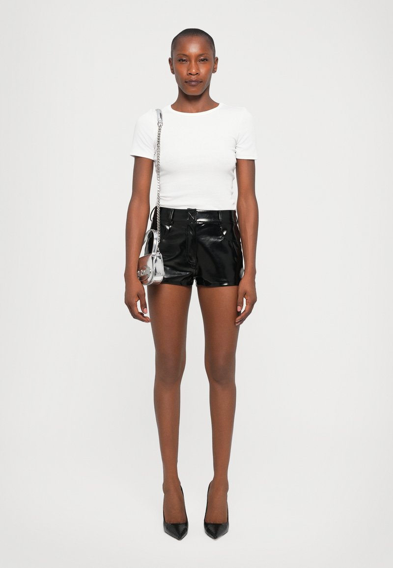 Woman wearing a white short-sleeve top, black shiny leather shorts, black heels, and a small metallic silver chain purse with a round design.