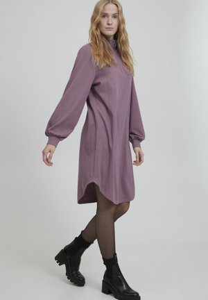 Lila Kleid mit langen, puffigen Ärmeln und hohem Kragen. Waffelstruktur und abgerundeter Saum. Schwarze Knöchelstiefel mit dicken Sohlen.