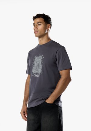 Jeune homme aux cheveux courts et bouclés porte un t-shirt gris foncé avec un motif détaillé d'ours en peluche, debout avec une main dans la poche.