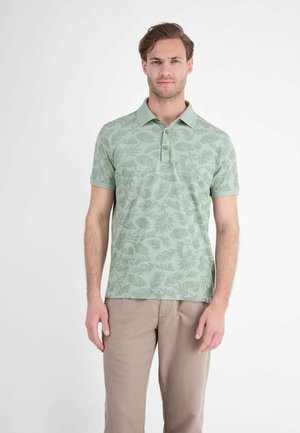 Man staat, draagt een lichtgroen poloshirt met bladpatronen en beige broek tegen een effen witte achtergrond.
