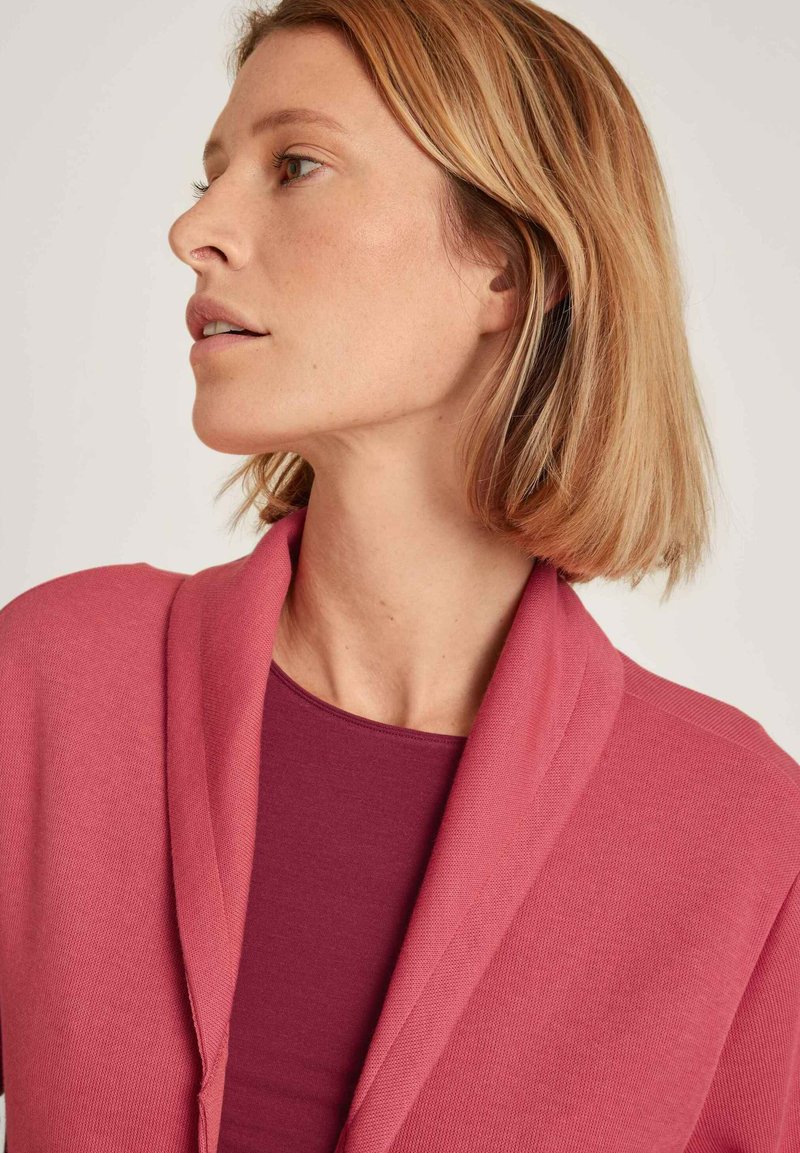 Rosa Schalkragen-Cardigan aus strukturiertem Strick, über einem tief burgunderroten, figurbetonten Oberteil mit glatter Oberfläche und entspannter Silhouette getragen.