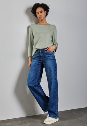 Hellgrüner, strukturierter Oberteil mit weitem Ausschnitt, kombiniert mit hochtaillierten blauen weit geschnittenen Jeans und weißen Sneakers. Stehend vor einer neutralen Wand.