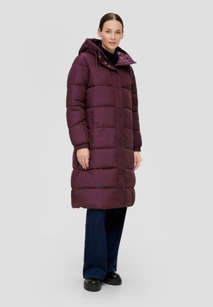Vrouw in een lange bordeauxrode pufferjas met capuchon, donkere broek en zwarte schoenen, staand tegen een effen lichte achtergrond.
