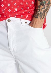 Pantaloni in denim bianco con tasca frontale, chiusura con bottone metallico e dettagli di cucitura discreti, abbinati a una camicia rossa con motivo.