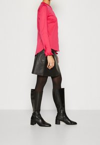 Mujer con una blusa de manga larga rosa brillante, falda de cuero negra, medias negras y botas de tacón altas negras, de pie de lado.