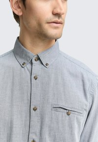 Chemise à motifs gris clair avec un col boutonné, texture subtile, poche poitrine et boutons foncés le long de la patte de boutonnage.