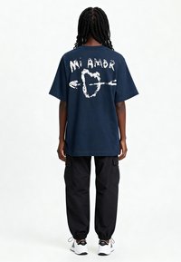 T-shirt en coton bleu marine avec un texte graphique blanc "Mi Amor" et un design de cœur au dos. Associée à un pantalon cargo noir et des chaussures de sport.