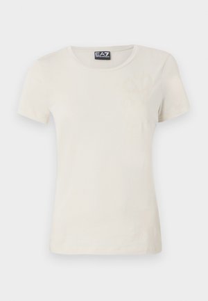 T-shirt beige clair à manches courtes en tissu doux, avec un col ras du cou et un motif subtil à l'avant près du coin supérieur gauche.