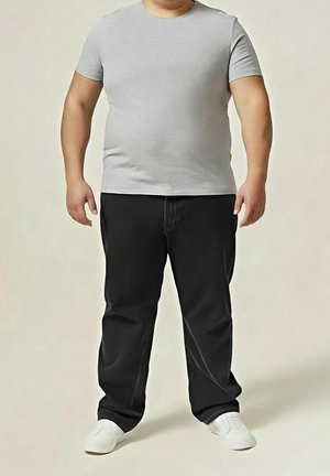 Homme portant une chemise cintrée à manches courtes gris clair, un pantalon noir ample et des chaussures blanches, debout, les bras détendus le long du corps.