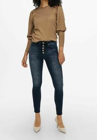 Pull en maille marron avec manches bouffantes, assorti à un jean bleu foncé taille haute avec une fermeture à cinq boutons et des effets d'usure légers, complété par des talons beiges.