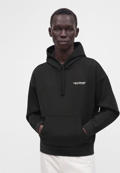 Άνδρας που φοράει μαύρο φούτερ Polo Sport Ralph Lauren με τα χέρια στην μπροστινή τσέπη, μπροστά από απλό ανοιχτό γκρι φόντο.