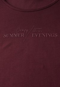 Camiseta de manga corta de color marrón con cuello redondo. Presenta un texto en relieve que dice "CÓMODAS NOCHES DE VERANO" con un texto adicional más pequeño debajo. Textura suave.