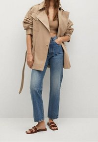 Femme portant une veste beige surdimensionnée, un haut court beige, un jean taille haute bleu et des sandales plates marron, se tenant devant un fond uni.