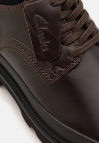 Barna bőrcipő sima felülettel, steppelt mintázattal, texturált rátéttel a "Clarks" logóval, valamint barna fűződíszítéssel.
