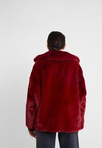 Röd konstpälsjacka med bred krage, med en mjuk textur och lös passform. Synlig sömnad längs kanterna. Bärs över mörka jeans.