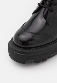 Botte en cuir noir poli avec semelle épaisse en caoutchouc texturé et bout arrondi, avec coutures visibles et lacets noirs.