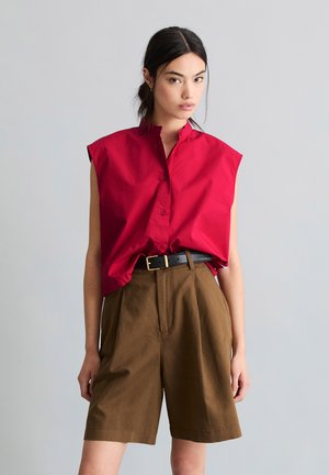 Kobieta ubrana w czerwoną koszulę bez rękawów zapinaną na guziki, wpuszczoną w brązowe szorty z wysokim stanem i czarnym paskiem, stoi na jednolitym tle.